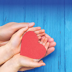 Picture of adult hands, cupping the hands of a child holding a red heart, on a blue table.